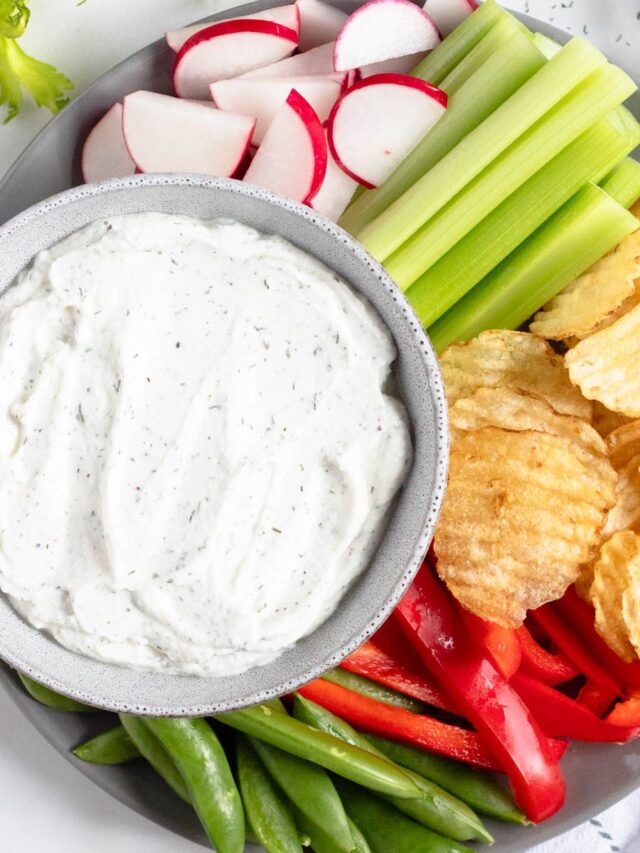 A bowl of cottage cheese dip surrounded by dippers like potato chips, celery sticks, red peppers, snap peas, and radishes.