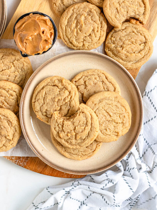 Top view of a plate of peanut butter cookies next to the remaining cookies cooling on parchment paper with a small dish of creamy peanut butter alongside.